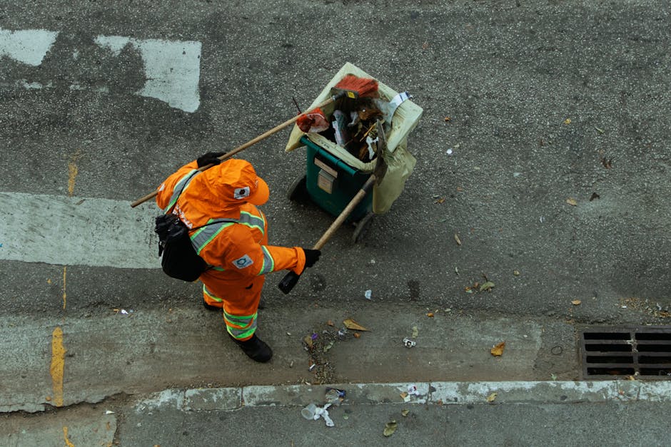 A street cleaner dressed in bright orange high-visibility clothing and a matching cap is sweeping debris from the edge of a sidewalk with a long-handled broom. The cleaner is carrying a black backpack-style bag and is working near a small waste collection bin containing various trash items. The surface is a textured asphalt road with scattered litter, debris, and a few fallen leaves, alongside a concrete curb with some dirt and small trash pieces. The area is illuminated by natural daylight, casting soft shadows, and the scene is part of a pavement cleaning operation. Cleaner Pimlico is providing surface cleaning and waste management services to maintain hygiene and cleanliness in the Pimlico area, aligned with the topic of bulking waste disposal versus professional cleaning.