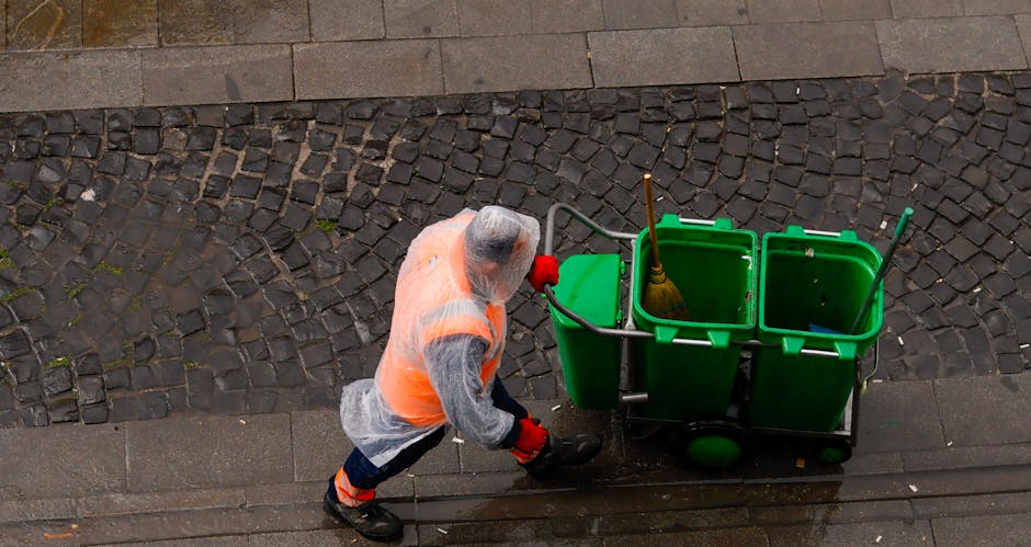 A street cleaner from Cleaner Pimlico, wearing a high-visibility orange vest, grey shirt, and red gloves, is pushing a green three-compartment cleaning trolley with a yellow broom and mop inside. The cleaner is walking on a concrete pavement next to a cobblestone street, with the trolley positioned beside them. The surrounding area appears wet, with some water visible on the pavement, indicating ongoing cleaning or recent cleaning activity. Bright natural lighting illuminates the scene, highlighting the cleanliness of the surfaces and the organized tools within the trolley, emphasizing professional surface cleaning and maintenance services in a residential or commercial Pimlico setting.