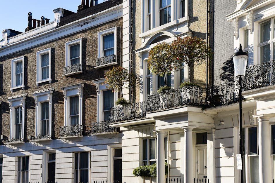 View of a modern multi-storey residential building with large glass windows and balconies, framed by leafy green tree branches in the foreground. The building features a combination of dark metal and 