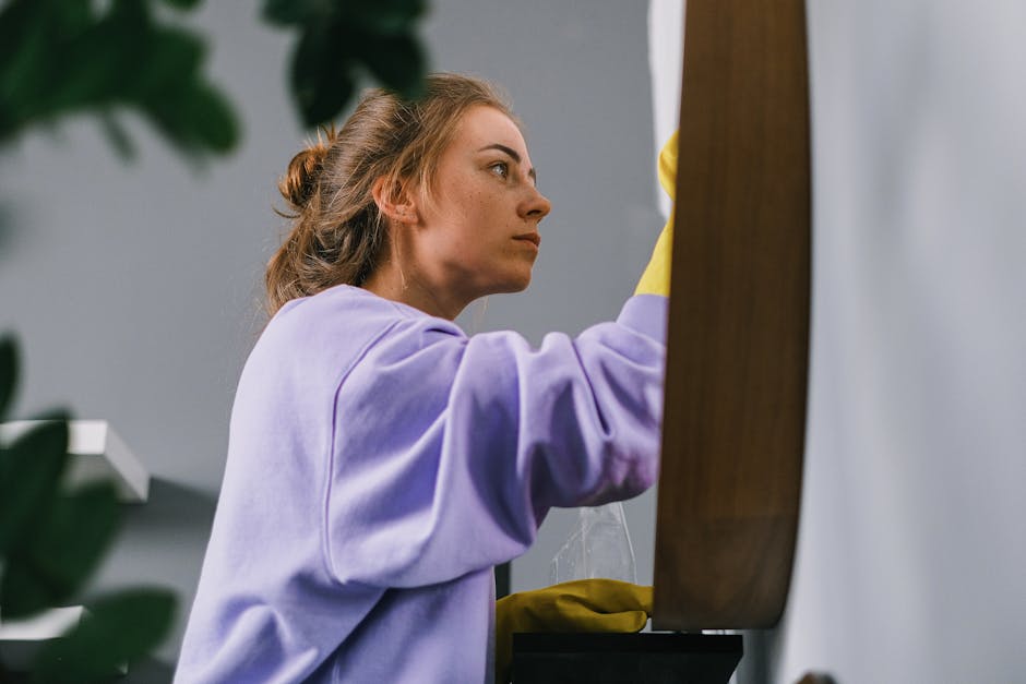 A young woman with light brown hair tied back, wearing a lavender sweatshirt and yellow cleaning gloves, is engaged in surface cleaning inside a residential or commercial space. She is carefully wiping a wooden surface, possibly a table or countertop, with a cloth, ensuring its cleanliness and shine. Bright, natural light illuminates the area, highlighting the smooth, polished texture of the wood. In the background, there is a grey wall and some green foliage, suggesting a tidy, well-maintained environment. The scene embodies professional deep cleaning and hygiene practices, aligning with [PAGE_TITLE], and exemplifies the quality cleaning services provided by [COMPANY_NAME].