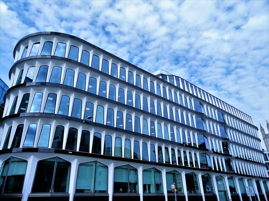 An outdoor urban scene featuring modern high-rise office buildings with extensive glass facades reflecting the blue sky. The foreground shows a curved, tiered amphitheater-style seating area made of c