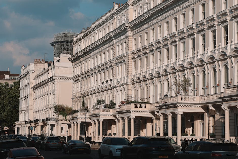 Image of a row of elegant, white Victorian-style terraced houses with ornate architectural details, large bay windows, and decorative columns along the ground floor. The buildings are well-maintained, with some balconies featuring potted plants. The street in front shows parked cars, and the scene is illuminated by natural daylight under a partly cloudy sky. This setting exemplifies an urban residential area where professional cleaning services by Cleaner Pimlico may be required for surface cleaning, deep cleaning, or maintenance to uphold hygiene and aesthetic appeal in properties similar to these flats.