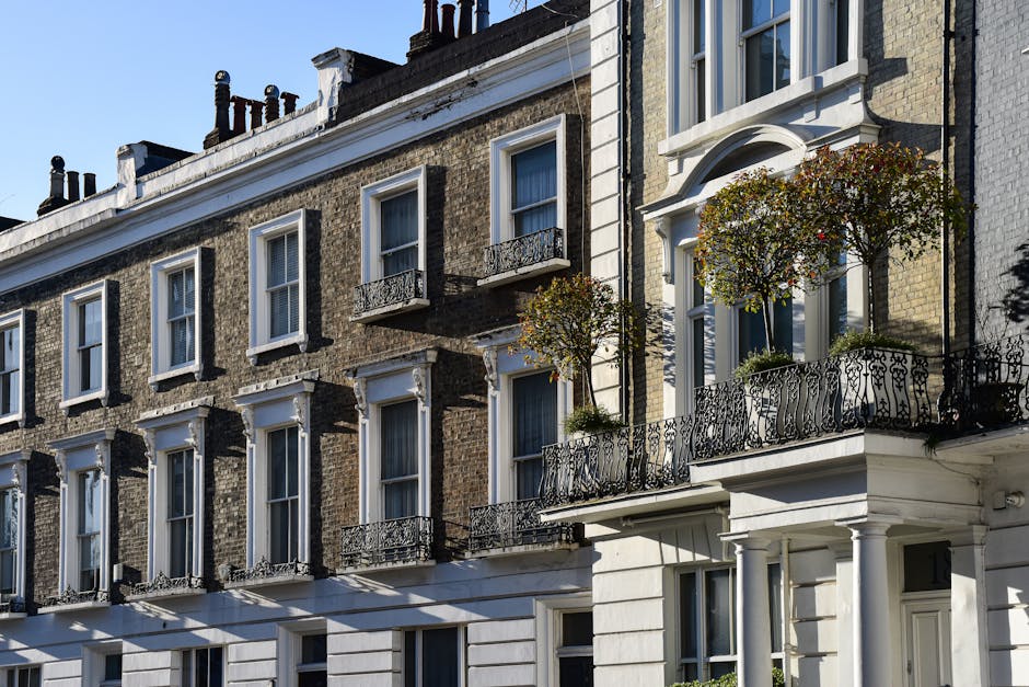 Exterior view of a traditional row of Victorian-style residential flats in Pimlico, featuring brick facades with white decorative trim and tall sash windows. The buildings have ornate black wrought iron balconies adorned with small trees and potted plants, creating a classic London street scene during daylight. The sunlight highlights the intricate architectural details and casts shadows on the light-colored stone base of the buildings, emphasizing their historic charm. Cleaner Pimlico provides professional cleaning services to maintain the hygiene and appearance of such residential properties, ensuring surfaces remain spotless and well-maintained for residents.