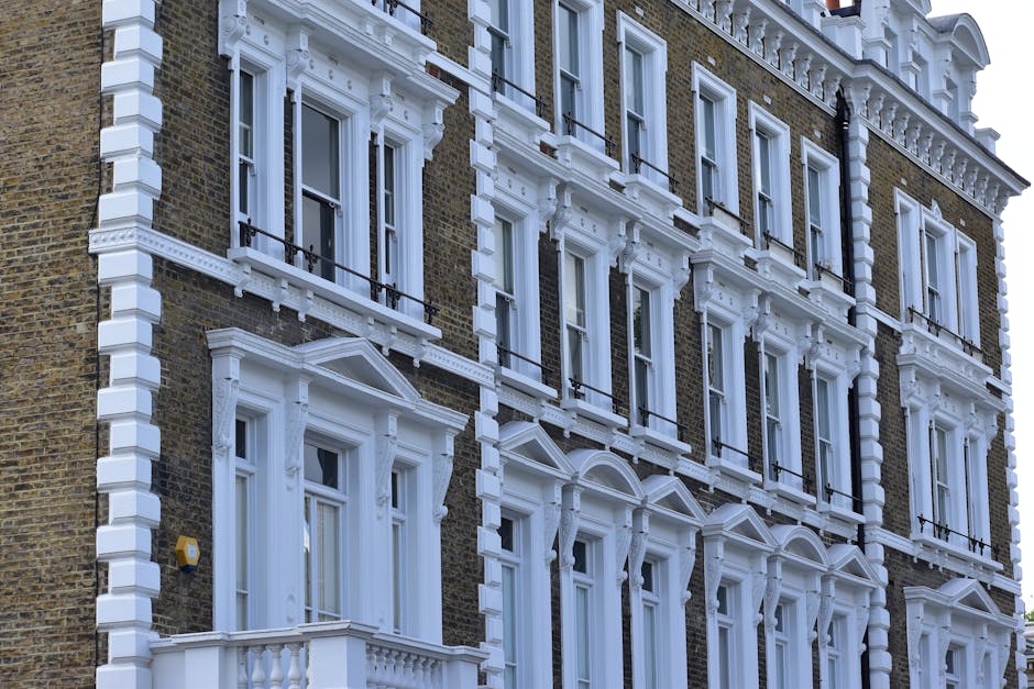Image of a row of elegant, white Victorian-style terraced houses with ornate architectural details, large bay windows, and decorative columns along the ground floor. The buildings are well-maintained, with some balconies featuring potted plants. The street in front shows parked cars, and the scene is illuminated by natural daylight under a partly cloudy sky. This setting exemplifies an urban residential area where professional cleaning services by Cleaner Pimlico may be required for surface cleaning, deep cleaning, or maintenance to uphold hygiene and aesthetic appeal in properties similar to these flats.