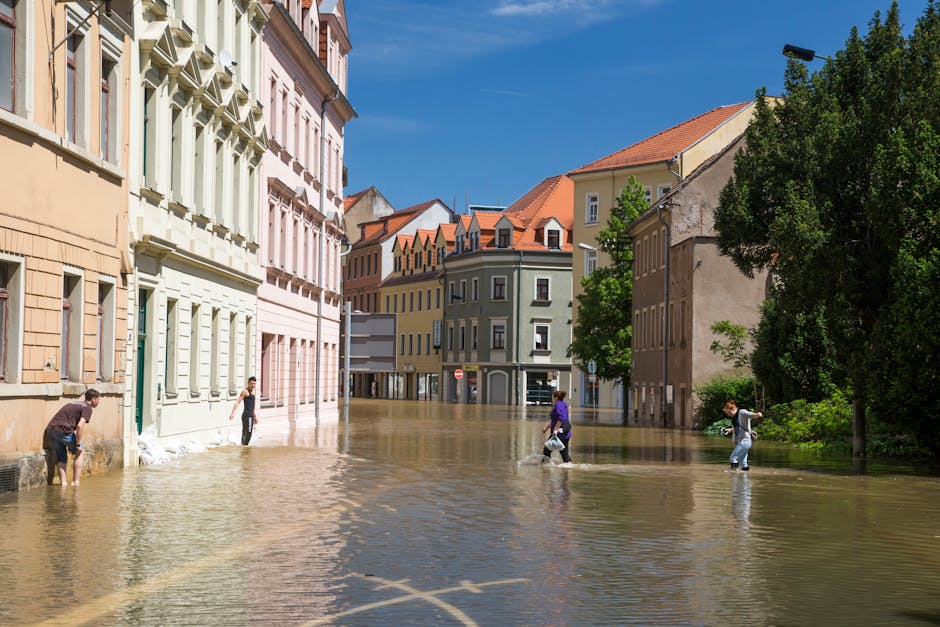 Photograph of a flooded urban street in Pimlico during daytime, with water level reaching halfway up the lower parts of multi-story buildings that feature pastel-colored facades and window shutters. Four pedestrians, dressed in casual clothing, wade through the floodwaters, some carrying bags or backpacks. The street is lined with trees and lampposts, with a clear blue sky overhead. The scene highlights the impact of recent flooding, emphasizing the importance of professional flood cleanup and surface sanitisation services, as offered by Cleaner Pimlico. The bright lighting shows the reflectiveness of the water on the pavement, and the buildings' clean, well-maintained exteriors contrast with the natural water flooding the street. This image illustrates the need for swift flood response and deep cleaning to restore safety and hygiene in affected areas.