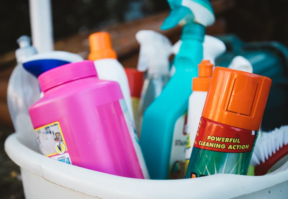 A collection of cleaning products in a white bucket, including a large pink bottle, a blue spray bottle, and an orange aerosol can labeled 'Powerful Cleaning Action,' along with other spray and pump bottles of various colors. The cleaning tools are arranged on a surface with a blurred background that suggests an outdoor setting. The scene displays a range of materials such as plastic bottles and spray nozzles, with the products appearing clean and ready for domestic cleaning tasks. The image emphasizes surface cleaning and sanitisation processes, aligning with the deep clean checklist for Pimlico homes provided by Cleaner Pimlico at SW1V. The well-organised assortment highlights tools and solutions used for routine and intensive cleaning in residential environments.