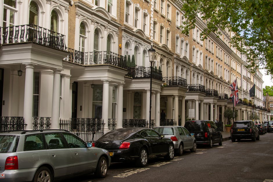 A row of elegant Victorian-style terraced houses in Pimlico, London, featuring white facades, tall windows, black wrought-iron railings, and small balconies. The street scene includes parked cars along the curb, with a mix of sedans and hatchbacks, and the pavement is clean with clearly marked parking spaces. A Union Jack flag and a black flag are visible on some of the houses' façades, and lush green trees line the street, providing natural shade. The lighting indicates a cloudy day, enhancing the clean and well-maintained appearance of the residential area, showcasing the classic architectural details typical of Pimlico. Cleaner Pimlico provides expert domestic and commercial cleaning services for maintaining such pristine environments.