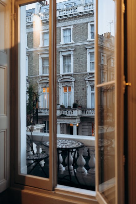 View through an open wooden-framed glass door onto a small balcony with a decorative black metal table and matching chairs. The exterior of an elegant, multi-story brick building with white decorative architectural details and tall windows is visible across the street. Warm interior lighting highlights the polished wooden door frame and the balcony's stone balustrade, creating a contrast between the indoor cleanliness and urban outdoor environment, indicative of deep surface cleaning and maintenance by Cleaner Pimlico for residential properties.