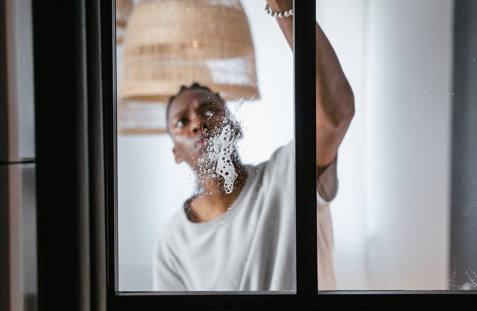 A woman with dark hair tied back, wearing a white t-shirt, is seen cleaning a glass window from inside a kitchen or living area. She is using a cloth or sponge to wipe condensation or dirt off the glass, with visible soap or cleaning solution bubbles. The window has a black frame, and behind it, part of a woven lampshade and a bright room with white or light-colored walls are visible, illuminated by natural light. The scene highlights surface cleaning and sanitisation practices as part of domestic cleaning services offered by Cleaner Pimlico in a residential setting.
