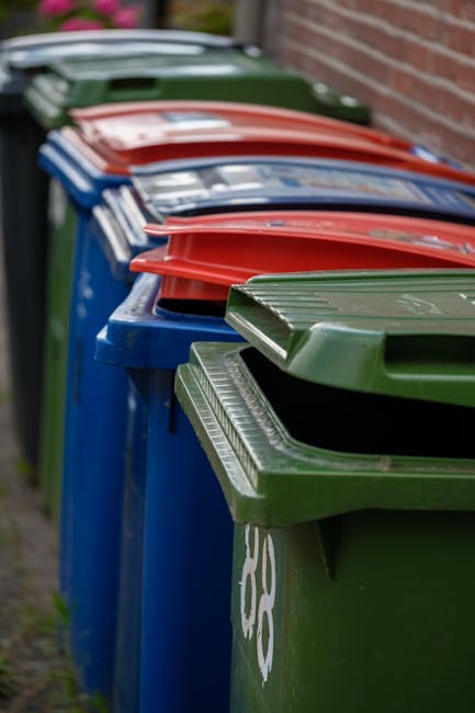 A row of outdoor refuse bins with colored lids—green, blue, red, and brown—lined up along a pavement next to a brick wall. The green bin in the foreground has a slightly open lid revealing some wa